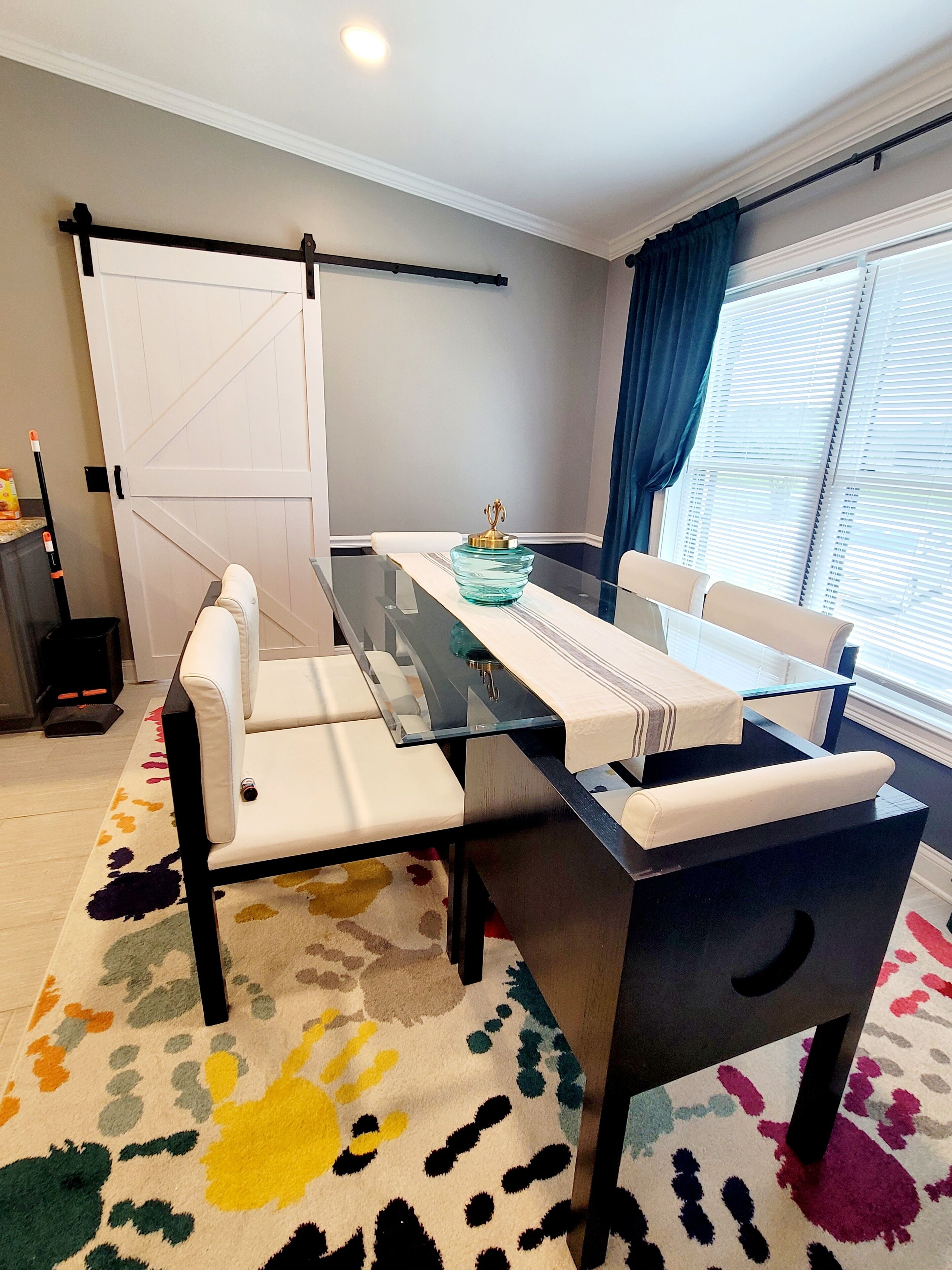 Un espace repas moderne avec une table à plateau en verre, des chaises blanches rembourrées, un tapis coloré et une fenêtre avec des stores.