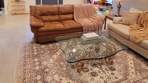Een ruime woonkamer met een bruine leren bank, een beige bank en een vloerkleed met een patroon. In het midden staat een glazen salontafel.