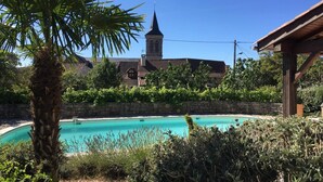 Una piscina con un muro de piedra y abundante vegetación en primer plano, un pérgola de madera, y un edificio con una torre al fondo.