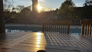 Una terraza de madera con un cuenco de comida, una cuchara y una taza, con vistas a un cuerpo de agua y un atardecer de fondo.