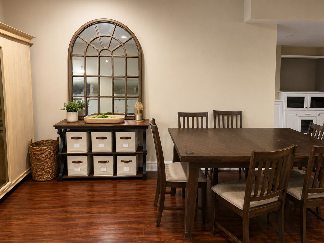 A dining room with a wooden table and chairs, a large arched mirror, and storage bins.