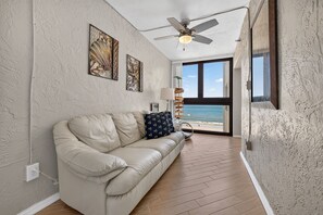A room with a beige leather sofa, a ceiling fan, and artwork on the walls. - Oceanfront Bliss – 2BR/2BA Condo with Stunning Views @ Sunrise (New Smyrna Beach)