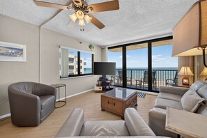 A modern living room with a ceiling fan, a flat-screen TV, and a view of the ocean. - Oceanfront Bliss – 2BR/2BA Condo with Stunning Views @ Sunrise (New Smyrna Beach)