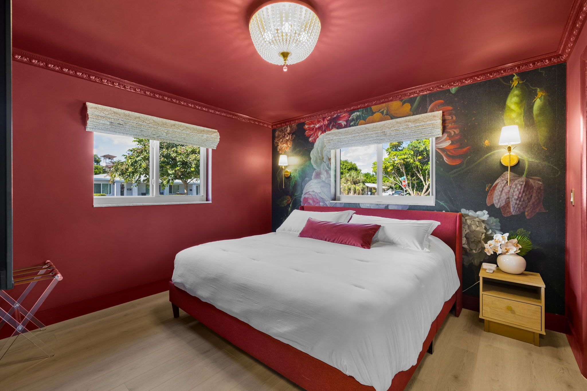 A bedroom with a red wall, a large bed, a chandelier, two windows with white curtains, and a nightstand with a lamp.