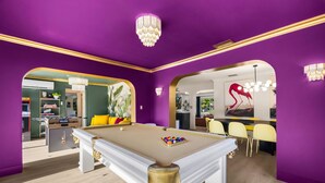 Ein Billardtisch in einem Raum mit violetten Wänden und goldenen Akzenten, ein Kicker und ein Essbereich mit gelben Stühlen.