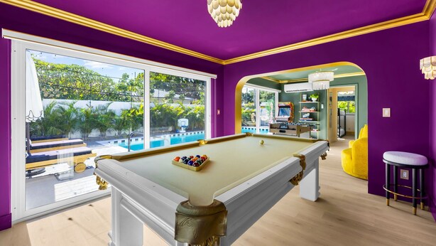 Ein Billardtisch in einem Raum mit violetten Wänden und einem großen Fenster mit Blick auf einen Pool und Grünflächen.