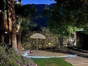 A nighttime outdoor area with a patterned umbrella, a stone bench, and a pathway. - Stylish Chic Escape, Private Pool & Golf Course Residence. Fun & Fabulousity (Palm Springs)