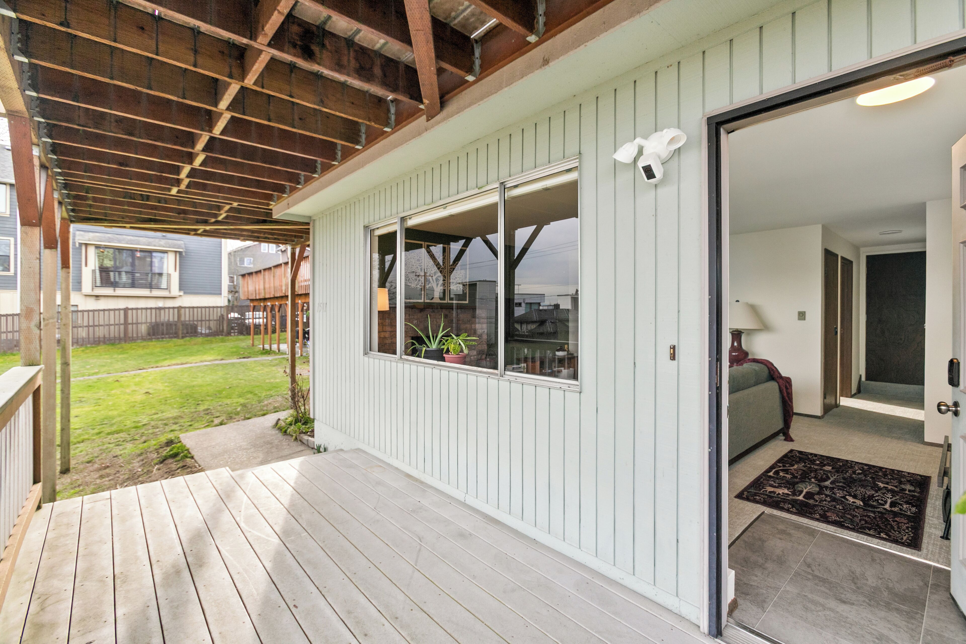 A covered porch with a wooden deck, a sliding glass door leading to an interior room with a sofa and a lamp, and a view of a backyard with a lawn and a fence.
