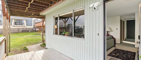 A covered porch with a wooden deck, a sliding glass door leading to an interior room with a sofa and a lamp, and a view of a backyard with a lawn and a fence.