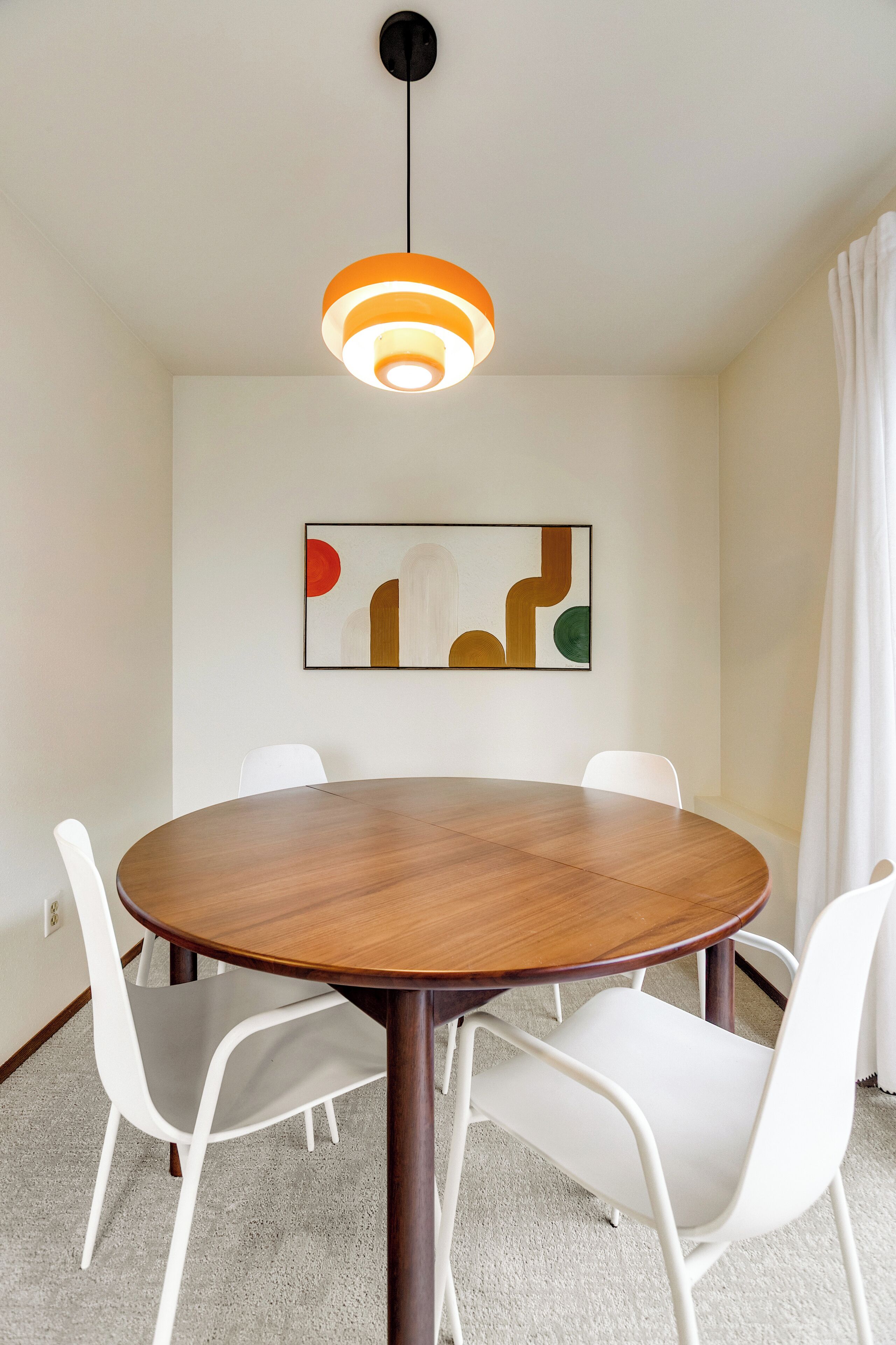 Un comedor con una mesa redonda de madera, sillas blancas, una lámpara colgante moderna y una obra de arte abstracto en la pared.