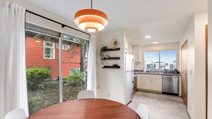 Een moderne keuken met witte kasten en roestvrijstalen apparatuur, een eethoek met een houten tafel en witte stoelen, en een groot raam met uitzicht op een rood gebouw en groen buiten.