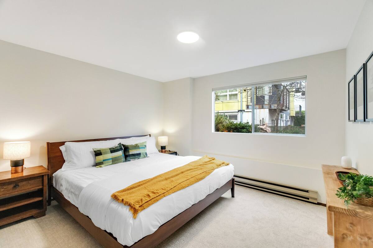 Une chambre à coucher avec un grand lit, des lampes de chevet, une fenêtre donnant sur des immeubles et une petite table de chevet en bois.