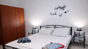 Una cama bien hecha con almohadas, un edredón doblado y toallas. Hay dos mesitas de noche con lámparas y libros. Un armario de madera está a la izquierda.