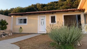 Una casa de un piso con techo de tejas, una puerta principal y dos ventanas. Un jardín con césped seco y un pequeño arbusto en el frente.