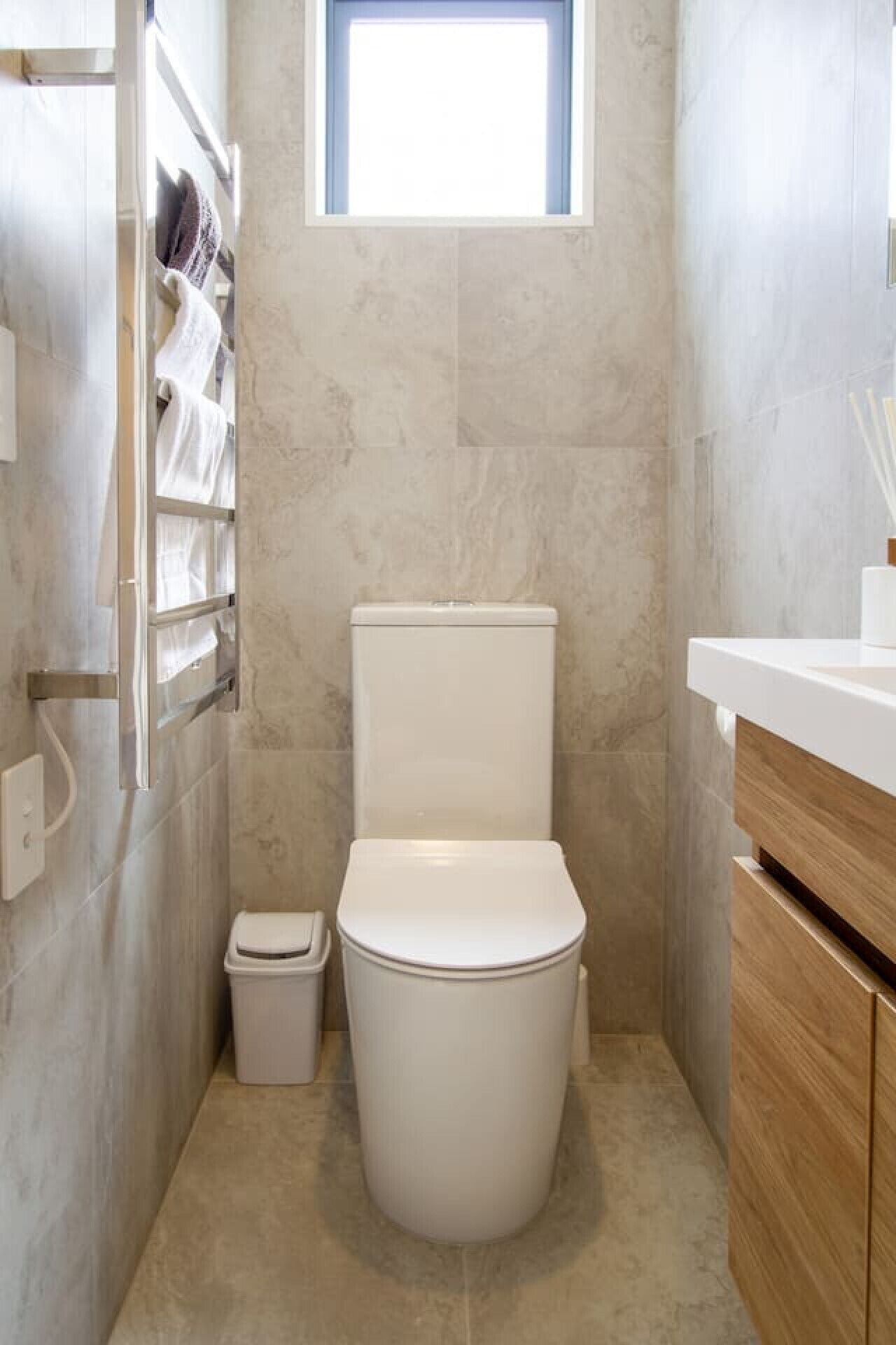 A compact bathroom with a toilet, a small trash bin, a window, and a wooden vanity with a white countertop.
