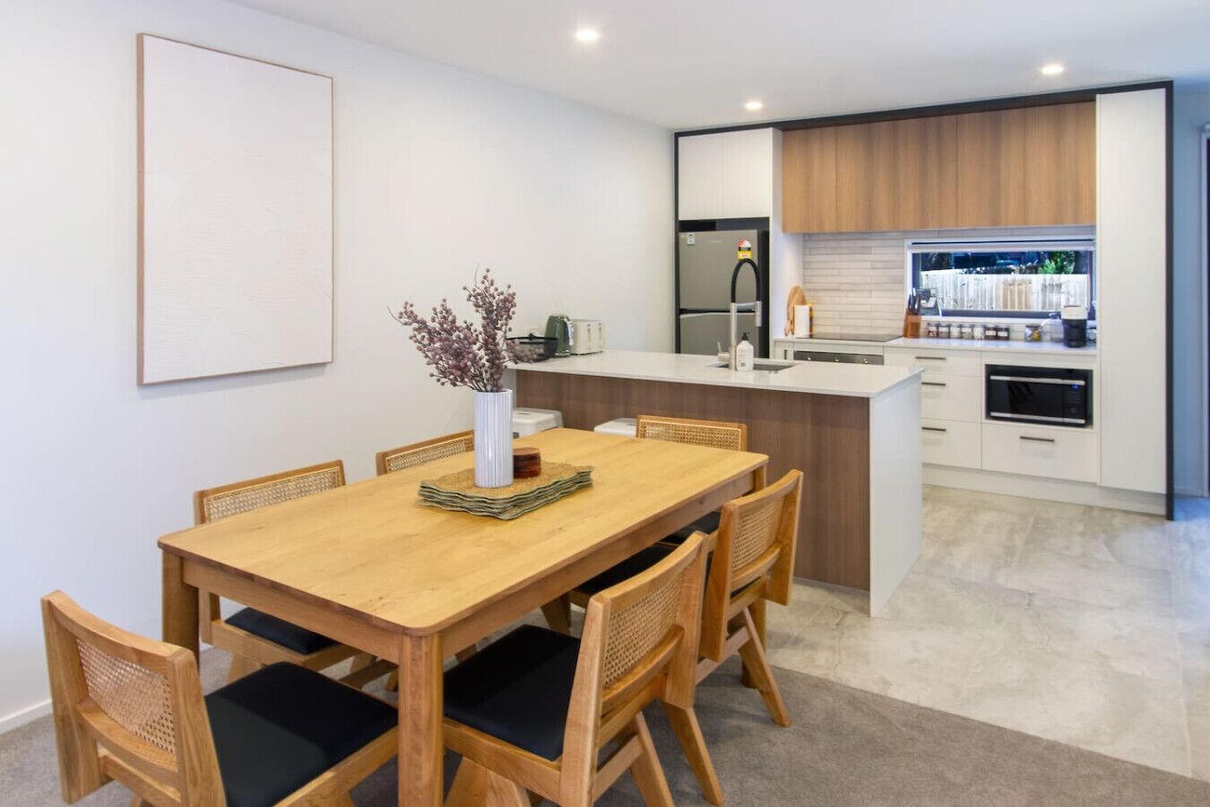 Una cocina moderna con una isla central, armarios blancos y un comedor con mesa y sillas de madera.