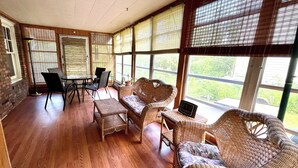 Ein Zimmer mit Korbmöbeln, einem Glastisch und großen Fenstern mit Jalousien.
