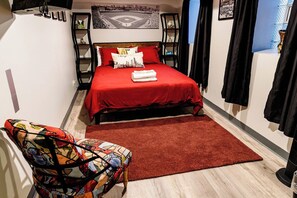 A hotel room with a bed, a chair, a red rug, and a cityscape painting on the wall.