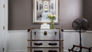 Une pièce avec un mur gris, un miroir reflétant un couloir, une valise ancienne dotée de tiroirs, un vase avec des fleurs blanches et un ventilateur sur pied.