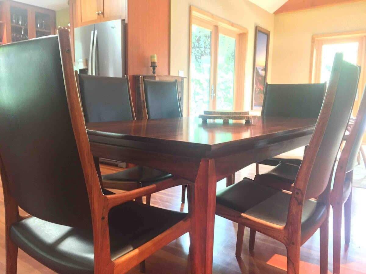 Un comedor con mesa y sillas de madera, un refrigerador y una ventana con vista a follaje.