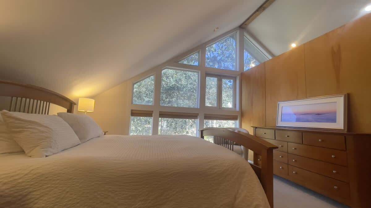 A bedroom with a large bed, a wooden dresser, and a large window with a view of trees.