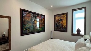 A bedroom with a large bed, two framed paintings, a window with a vase, and a nightstand with a lamp.