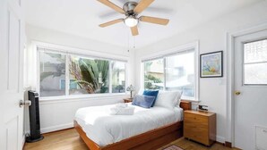 Un dormitorio con cama, mesitas de noche, ventilador de techo, calefactor portátil y ventanas amplias.
