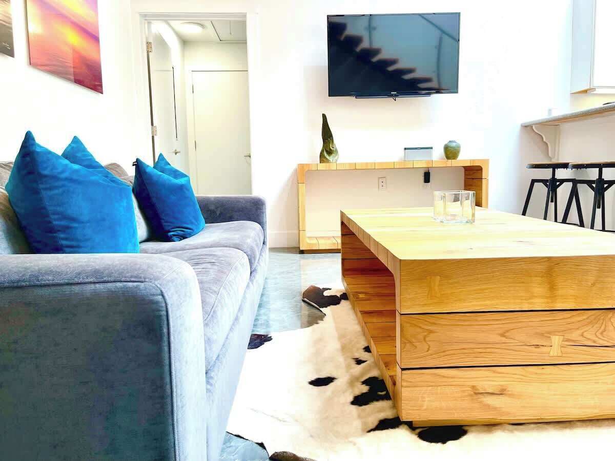 Een moderne woonkamer met een grijze bank, blauwe kussens, een houten salontafel, een tv aan de muur en een koehuidtapijt.