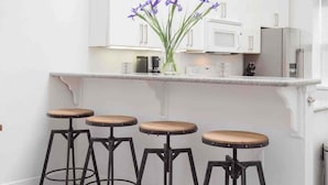 Una cucina con mobili bianchi, un forno a microonde e un frigorifero. Ci sono quattro sgabelli con sedute in legno e struttura in metallo nero. Sul bancone c'è un vaso con fiori viola.