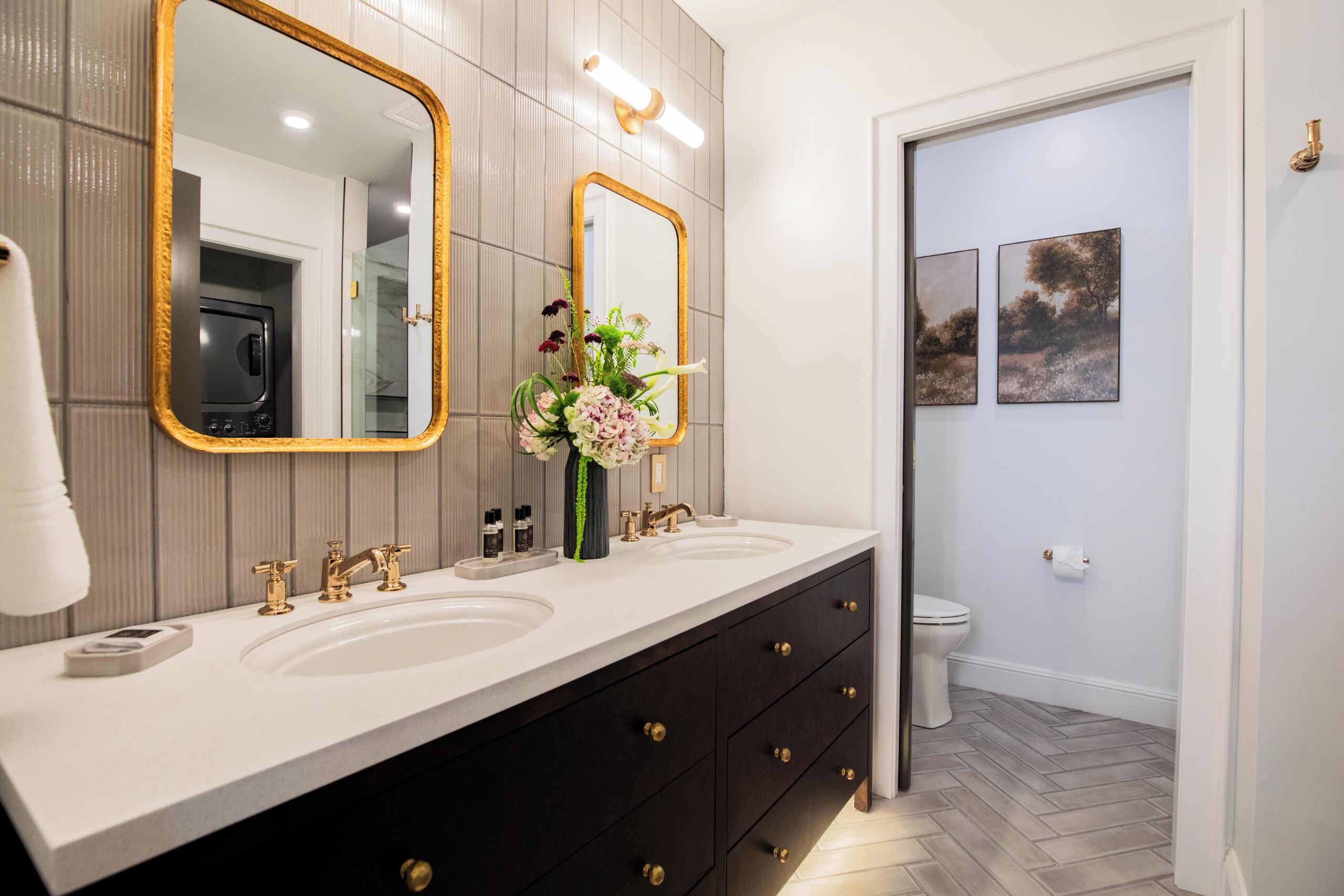 Ein Badezimmer mit einem Doppelwaschbecken, zwei Spiegeln, einer Blumenvase und einer Toilette.