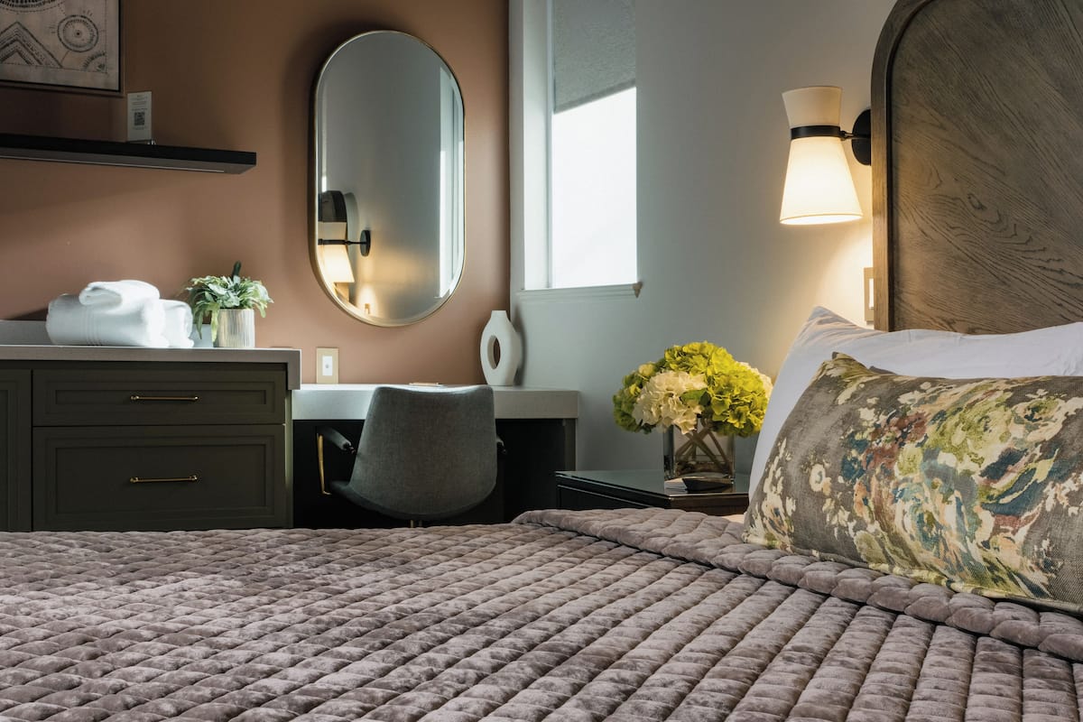 A hotel room with a bed, a desk, a chair, a mirror, a lamp, and a window.