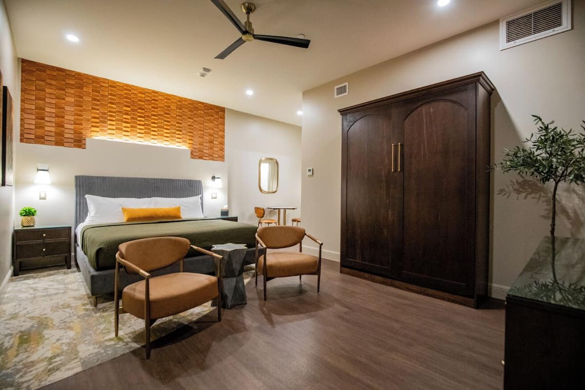 A bedroom with a bed, two chairs, a wardrobe, a ceiling fan, and a nightstand with a lamp.