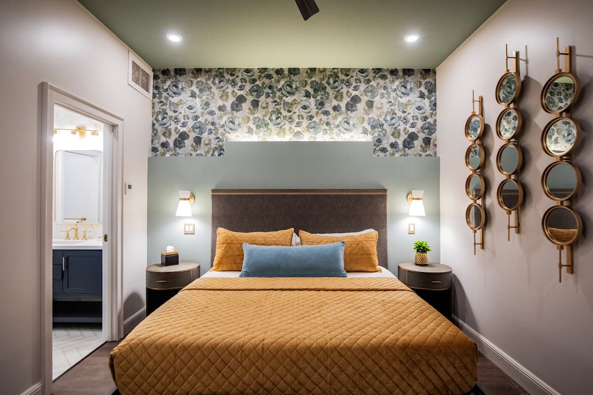 A bedroom with a bed, bedside tables, wall-mounted mirrors, a nightstand, a window with floral wallpaper, and a door leading to a bathroom.