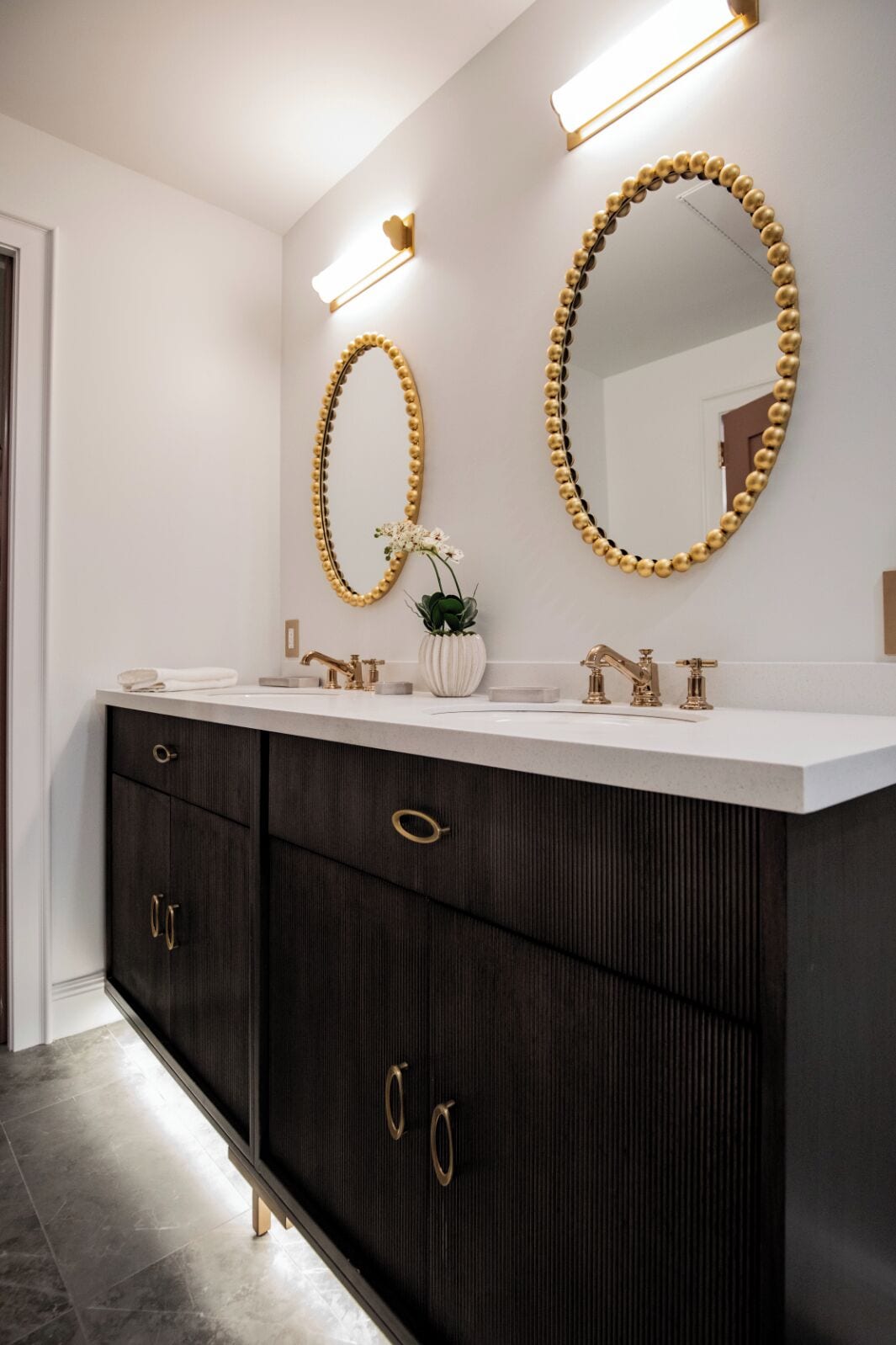 Un baño moderno con dos espejos grandes, una encimera blanca, muebles oscuros e instalaciones doradas.