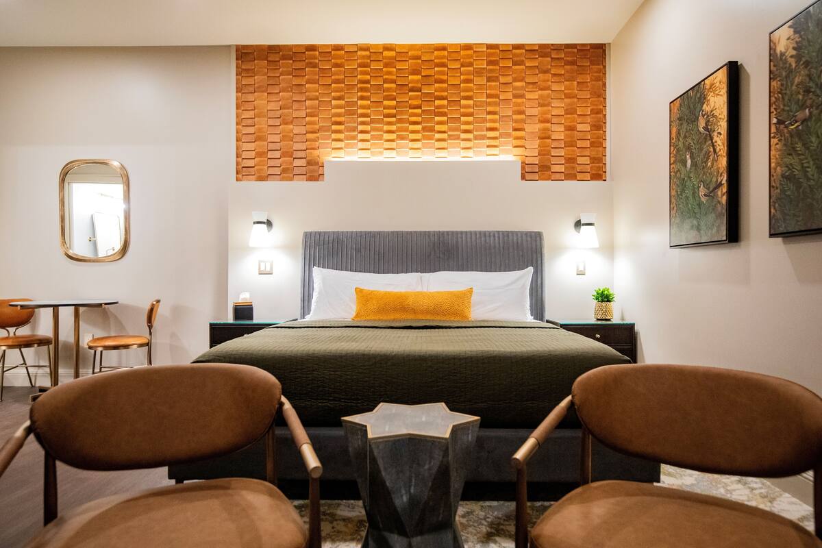 A modern bedroom with a brick-patterned headboard, a bed with a gray and yellow pillow, two brown chairs, a small table, and a mirror on the wall.