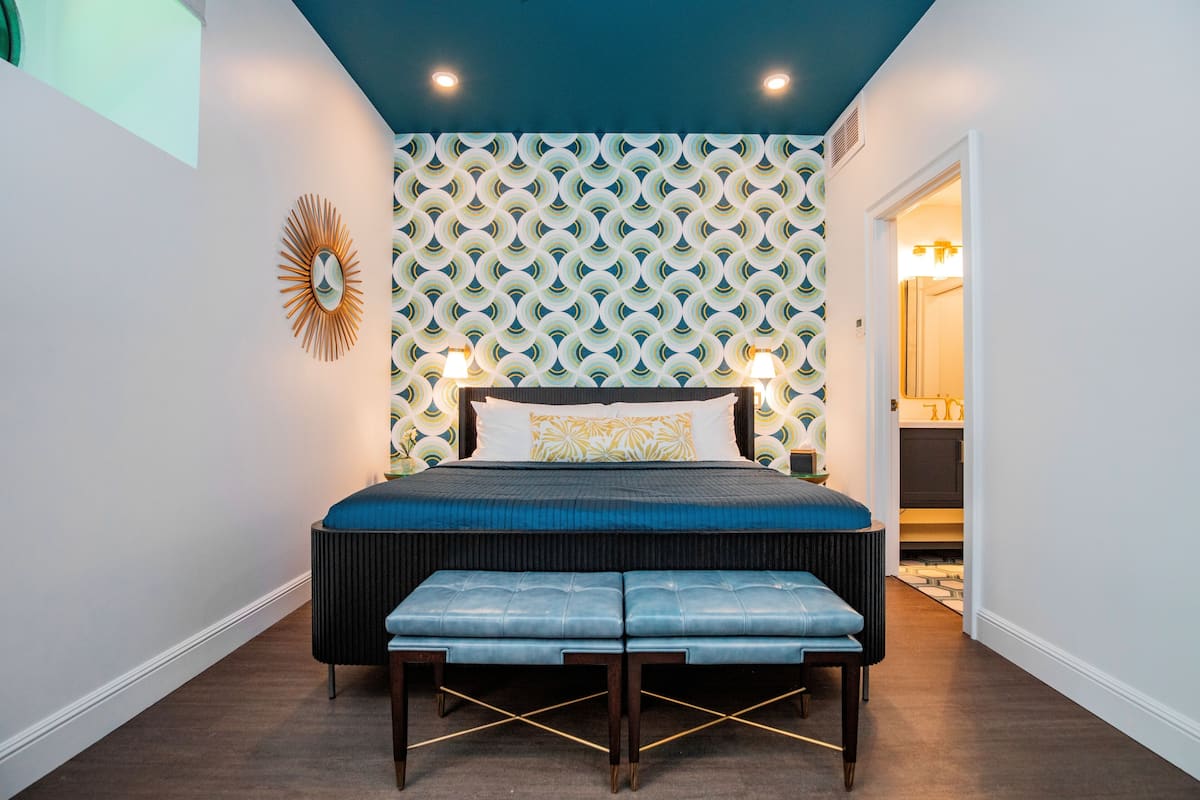 A bedroom with a patterned wall, a bed with a blue headboard, two bedside lamps, and a bench at the foot of the bed.