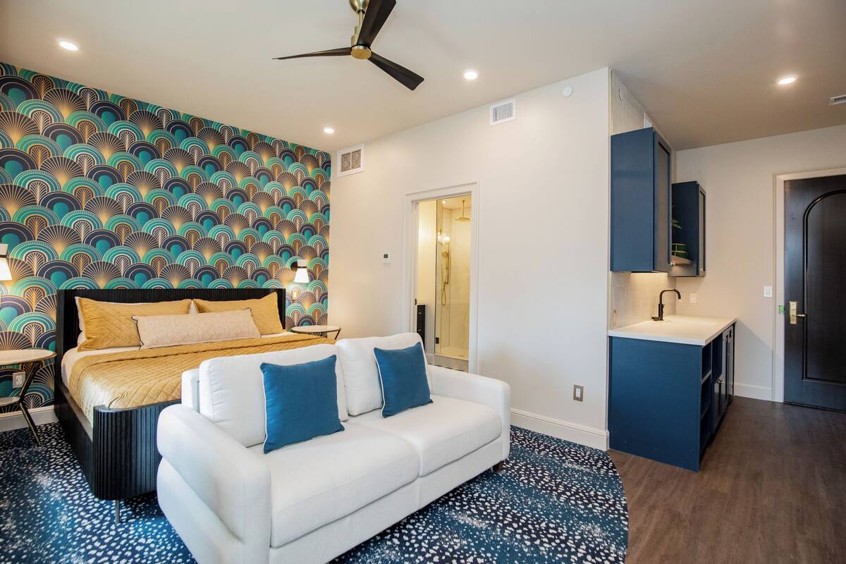 A bedroom with a bed, a white sofa, a ceiling fan, a bathroom with a shower, and a small kitchenette.