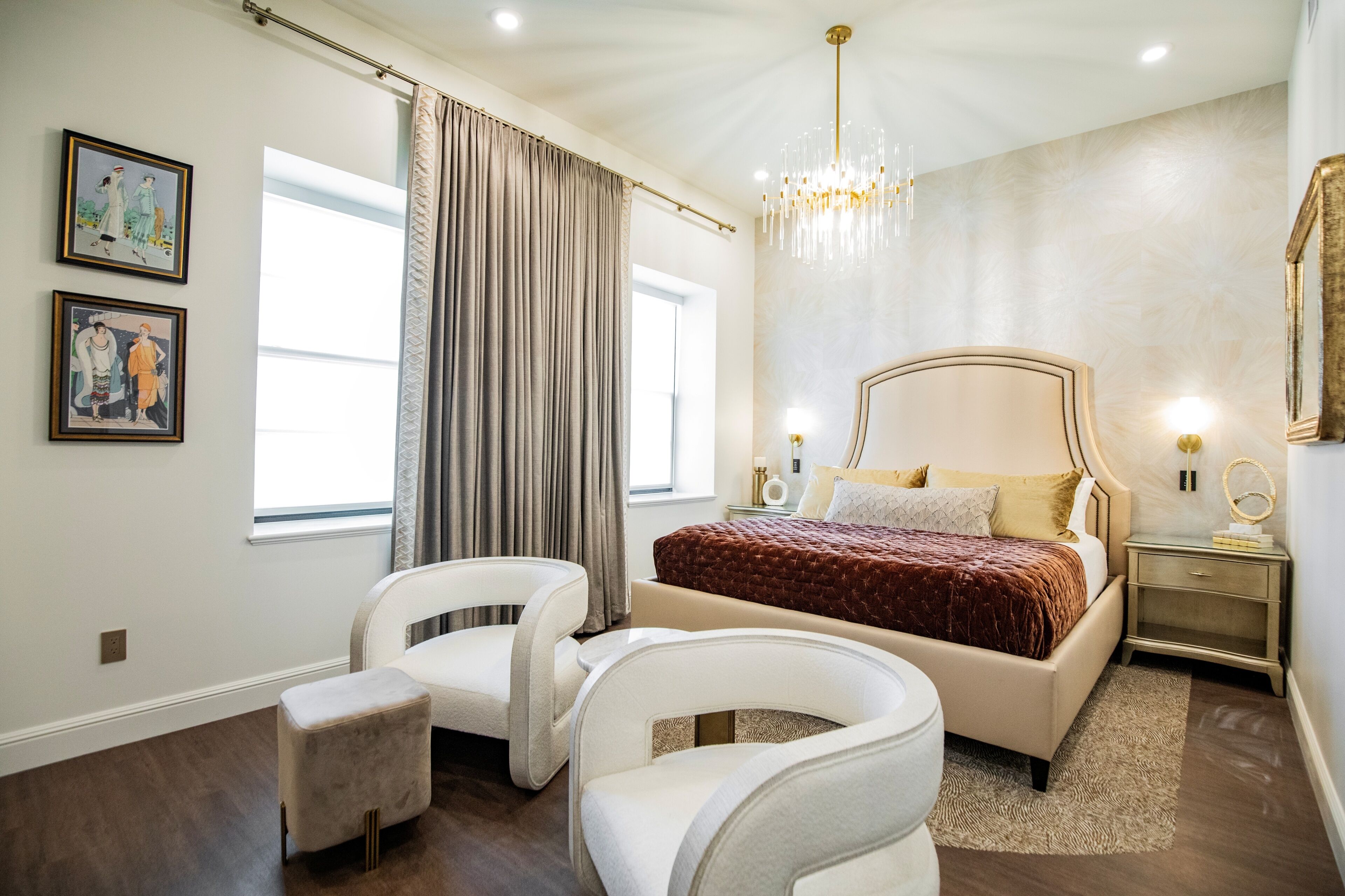 A bedroom with a bed, two chairs, a nightstand, a chandelier, and two wall-mounted lights.