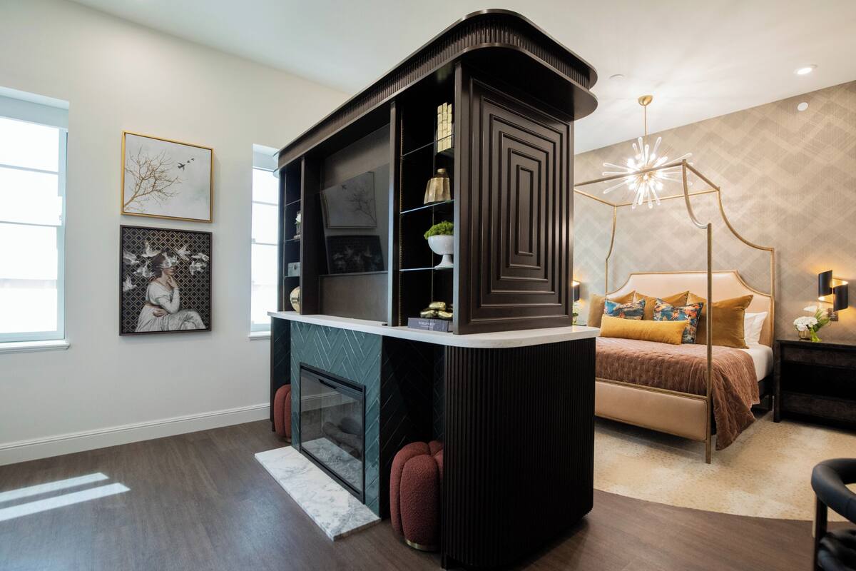 A modern bedroom with a built-in TV stand, a fireplace, a bed with a headboard, and a decorative ceiling light.