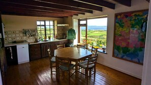 A kitchen with wooden flooring, a dining table with chairs, a view of the countryside through large windows, and a colorful painting on the wall.