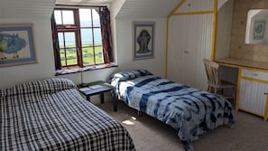 A bedroom with two beds, a desk, and a window with a view of the countryside.