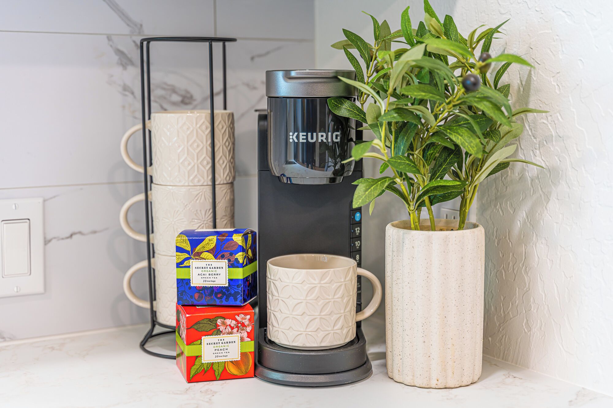 Un comptoir de cuisine avec une machine à café Keurig, deux boîtes de thé biologique, une tasse blanche et une plante en pot.