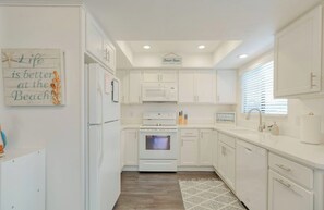 A kitchen with white cabinets, a microwave, oven, and a sink. There is a sign on the wall that says "Life is better at the Beach!" and another sign that says "Welcome to the Beach." - Modern Coastal Condo in Downtown Pismo Beach | Gated Parking | Walk to Beach (Pismo Beach)