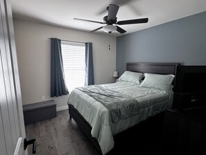 A bedroom with a bed, a ceiling fan, a window with curtains, a bedside lamp, and a nightstand. - Spacious 5-bedroom Pool Home near Beaches-Hot Tub, Kayaks, Massage Chair (North Port)