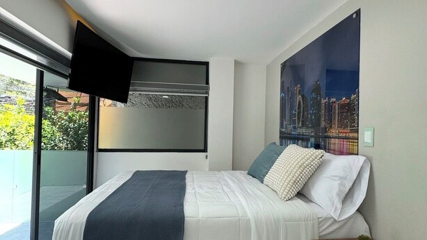 Un dormitorio moderno con cama, mesita de noche, televisor de pantalla plana en la pared y una pintura de paisaje urbano.