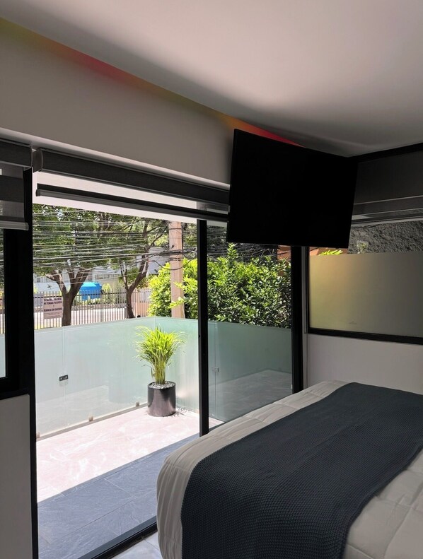 Une chambre moderne avec un grand lit, un téléviseur à écran plat fixé au mur et une porte-fenêtre coulissante donnant sur un balcon agrémenté d’une plante en pot.
