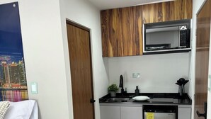 Cocina compacta con microondas, lavaplatos y fregadero. Hay una pequeña mesita de madera con una vela y una planta. La habitación tiene una cama blanca y vistas a un paisaje urbano a través de una ventana.