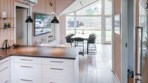 Une cuisine moderne dotée d’un grand îlot central, de meubles blancs et d’un coin repas avec une table et des chaises. Une grande fenêtre offre une vue sur l’extérieur.