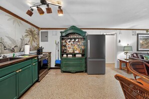 Une cuisine avec des meubles de couleur verte, un réfrigérateur, un coin repas avec des chaises en osier et un mur orné d’une fresque aux motifs tropicaux.