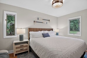 Une chambre avec un grand lit, deux tables de chevet, une petite table d’appoint, une lampe fixée au mur et deux fenêtres donnant sur des arbres.